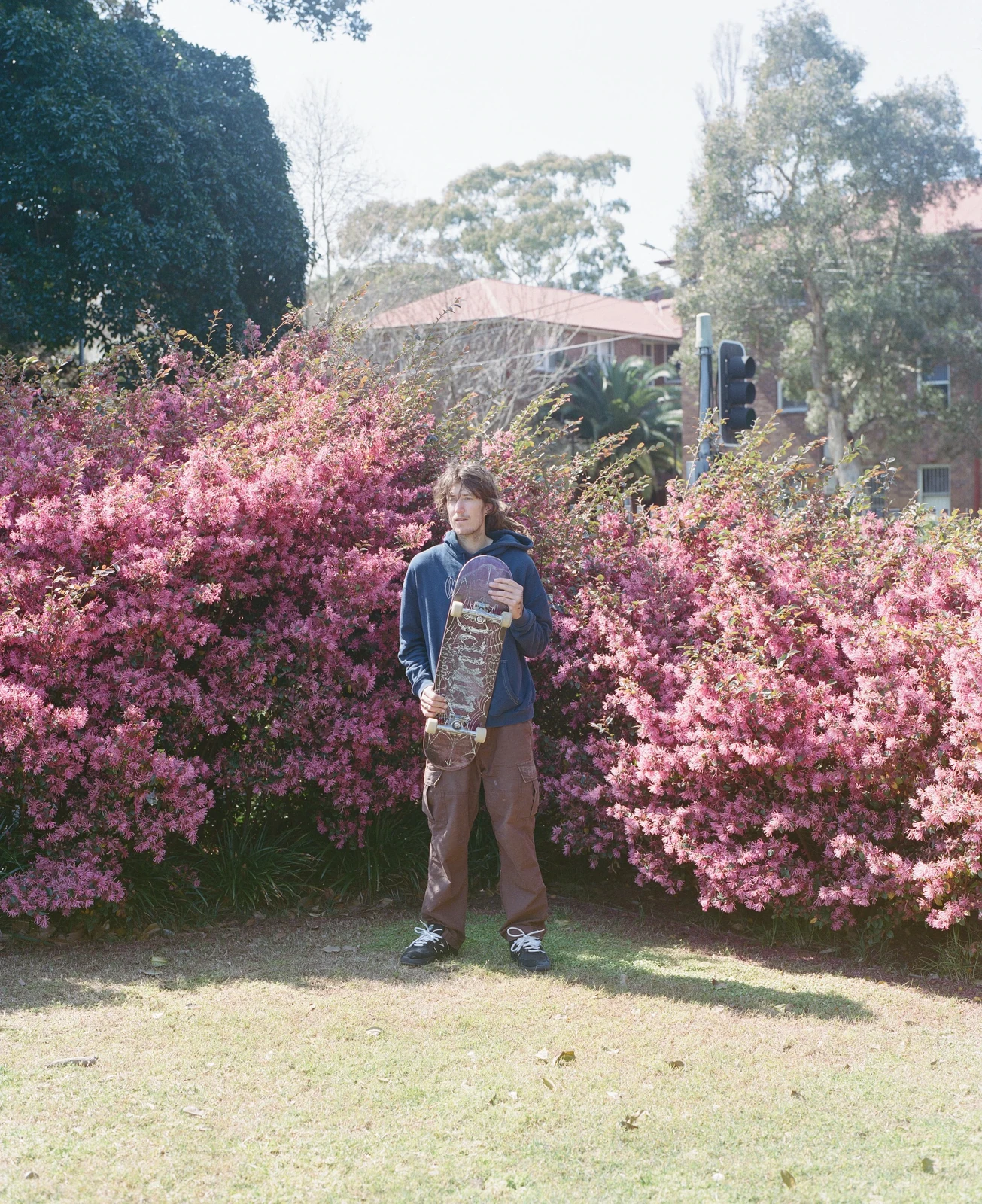 A young man standing in front of pink flowering bushes holding a skateboard.